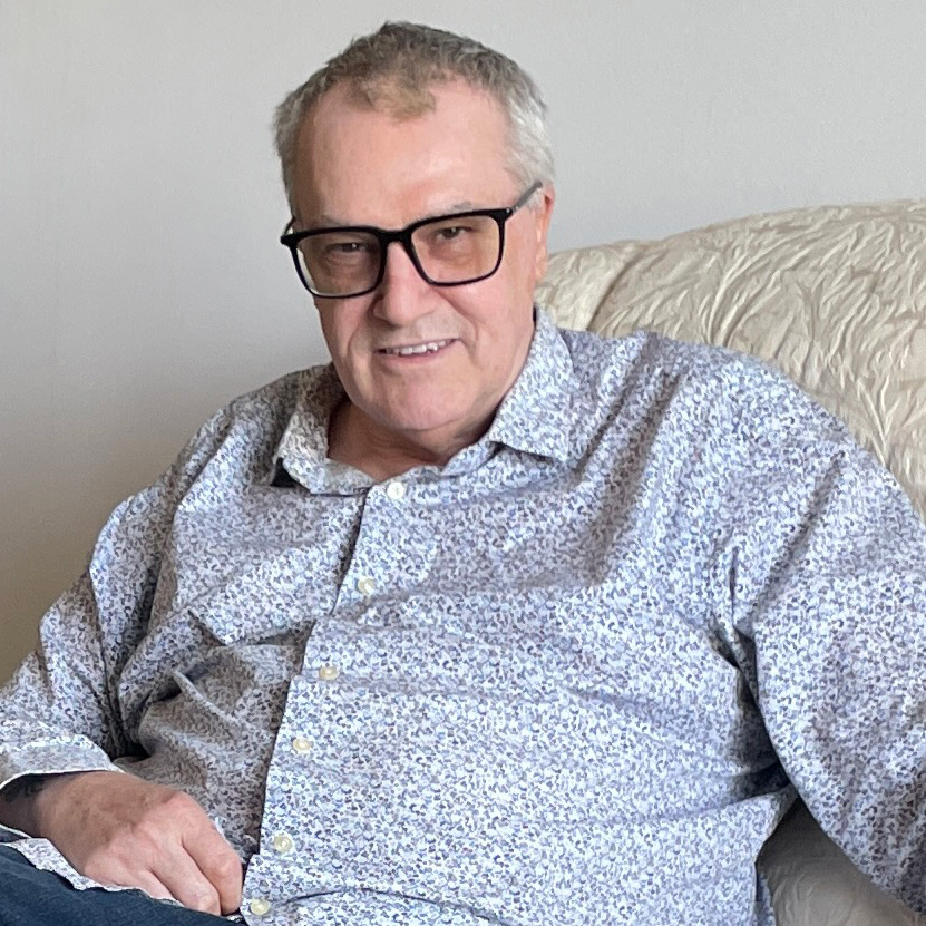 Un homme âgé et souriant portant des lunettes assis sur un canapé.