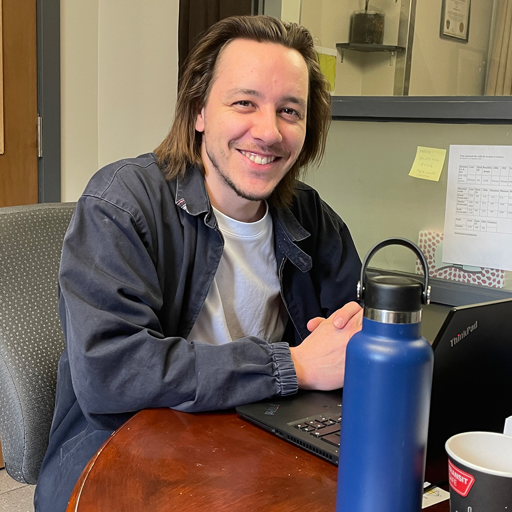 Un homme souriant à un bureau, travaillant sur un ordinateur portable avec une bouteille de café à côté de lui.