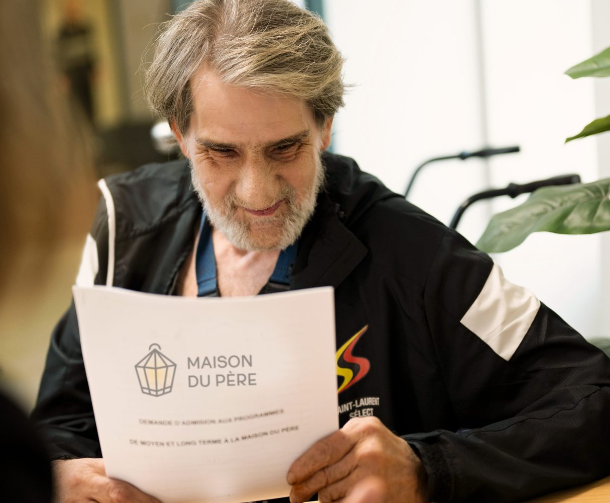 Un homme âgé souriant regarde un document ayant l'inscription La Maison du Père.