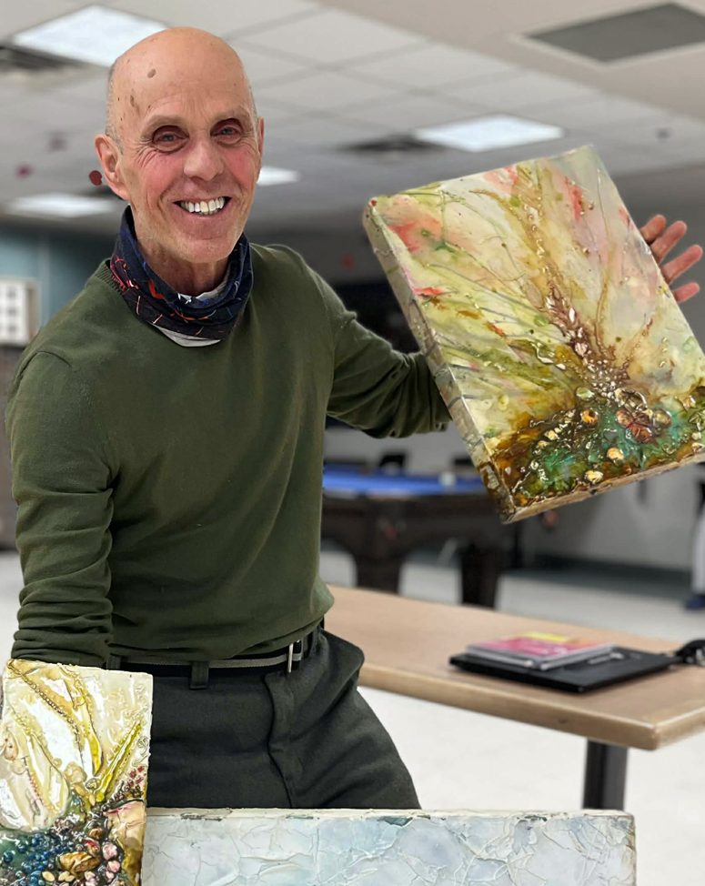 Un homme chauve et souriant tient une peinture dans une salle.