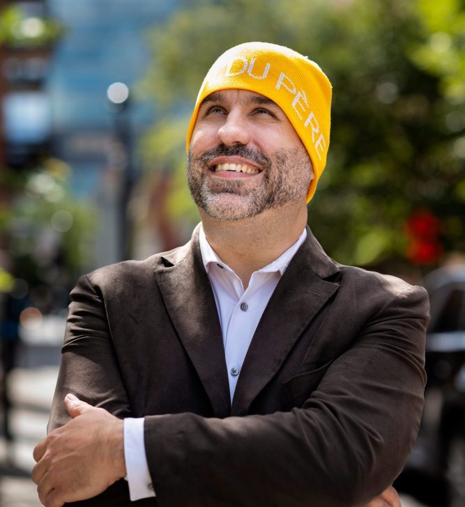 Un homme portant la tuque jaune de la Maison du Père sourie en regardant vers le ciel a les bras croisés.